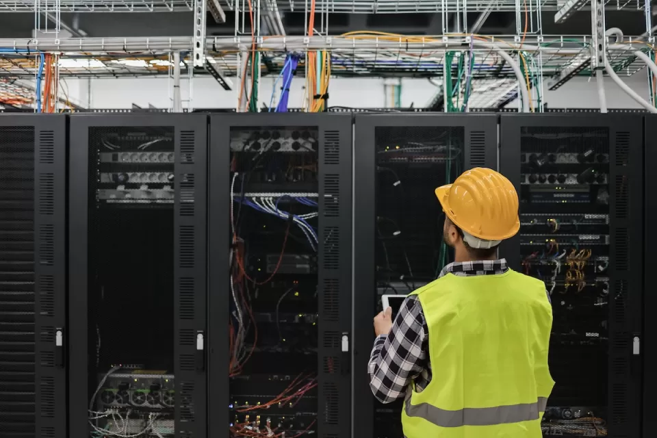 Worker in yellow safety vest and hard hat looks up at servers in data center.